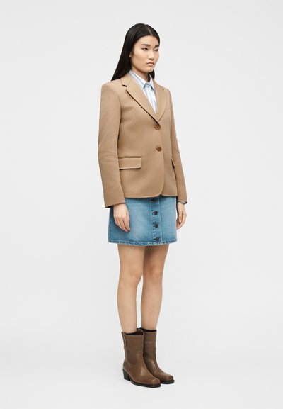 Γυναίκα που φοράει καφέ σακάκι, μπλε τζιν φούστα με κουμπιά μπροστά, ριγέ πουκάμισο και καφέ μπότες μέχρι τη μέση της γάμπας, στέκεται μπροστά σε ένα απλό λευκό φόντο.