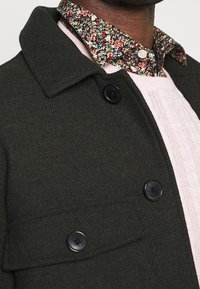 Dark green wool blend coat with a spread collar, featuring black buttons and a textured pink sweater beneath a patterned floral shirt.
