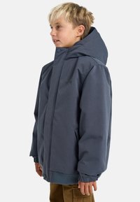 Ragazzo che indossa una giacca invernale grigio scuro con cappuccio e pantaloni marroni, in piedi di profilo su uno sfondo chiaro e semplice.