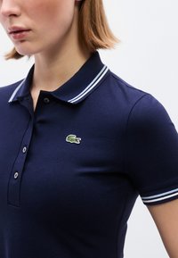 Femme portant un polo bleu marine avec des rayures blanches sur le col et les manches, orné d'un petit logo de crocodile vert sur la poitrine.