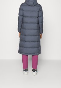 Long manteau rembourré gris foncé avec capuche, présentant un design matelassé et un léger éclat. Porté sur un pantalon rose et de grosses baskets blanches.