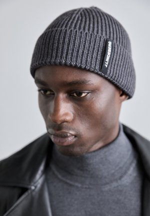 Bonnet en tricot côtelé gris avec un revers et une étiquette noire horizontale. Porté par une personne portant un col roulé gris et une veste noire.