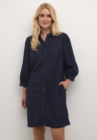 Robe bleu marine à boutons en tissu doux et texturé. Dotée d'un col, de manches bouffantes, de poches avant et d'accents en boutons marron.