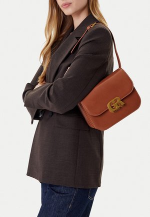 Frau trägt ein dunkelbraunes Blazer und blaue Jeans und hat eine kleine braune Leder-Umhängetasche mit goldener Schnalle am Arm.