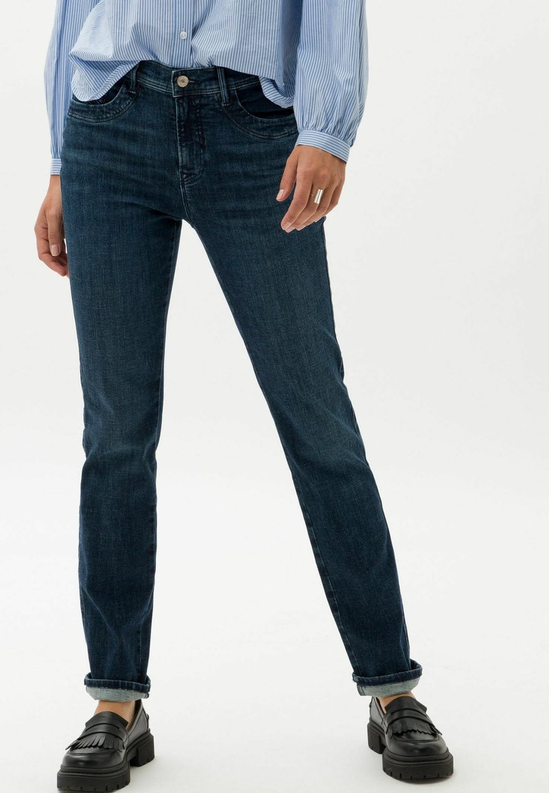 Personne portant un jean bleu foncé retroussé, des mocassins noirs à glands et une chemise à manches longues rayée bleu clair, les mains détendues sur les côtés.