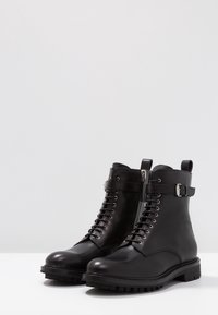 Botas de cuero negro hasta el tobillo con cordones en la parte frontal, cremallera lateral y un acento de correa con hebilla; suela de goma texturizada para tracción.