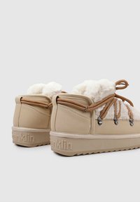 Zapatillas beige con forro de pelo, parte superior texturizada, cordones marrones, herrajes metálicos y logo "Franklin" grabado en la suela. Diseño de caña media.