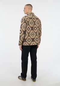 Man staat met zijn rug naar de camera, draagt een bruin en beige geometrisch patroonjacket, zwarte broek en donkergrijze sneakers met witte strepen.
