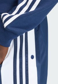 Tmavomodrá bežecká bunda s bielymi pruhmi, textúrovanou látkou a detailom so zapínaním na cvočky na boku, predstavujúca športový dizajn.