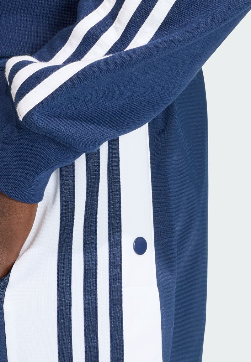 Marineblaue Trainingsjacke mit weißen Streifen, strukturierter Stoff und einem Druckknopfdetail an der Seite, das ein sportliches Design zeigt.