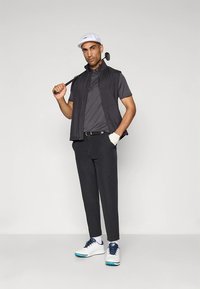 Golfový outfit zahŕňajúci tmavú bezrukávovú vestu, sivé polo tričko, čierne nohavice, biele topánky s modrými akcentmi a bielu čiapku.