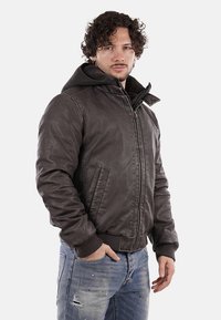 Giacca bomber in pelle marrone con cappuccio, chiusura con zip, finitura testurizzata, polsini e orlo a coste, indossata sopra jeans chiari strappati.
