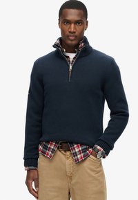 Marinblauer Strickpullover mit gerippten Bündchen und einem Reißverschlusskragen, über einem rot, grün und blau karierten Hemd getragen, kombiniert mit beigen Hosen.