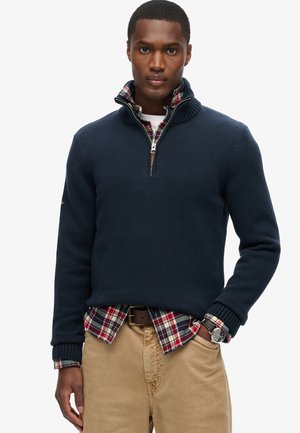 Marinblauer Strickpullover mit gerippten Bündchen und einem Reißverschlusskragen, über einem rot, grün und blau karierten Hemd getragen, kombiniert mit beigen Hosen.