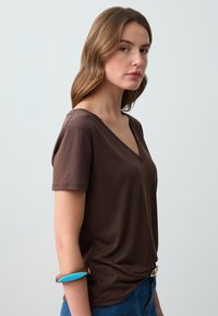 T-shirt marrone a scollo a V, maniche corte, tessuto morbido, vestibilità ampia, indossato con un bracciale blu. Esposto su un modello rivolto a sinistra contro uno sfondo chiaro.
