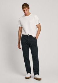 Valge lühikeste varrukatega T-särk, millel on tekst "Pure Jeans", kombineeritud tumedate siniste pükste ja valgete jooksujalanõudega, millel on pruunid tald, seisab heledal taustal.