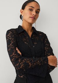 Schwarze Spitzenbluse mit klassischem Kragen, durchgehender Knopfleiste und Glockenärmeln mit Blumenmuster und transparenten Details.