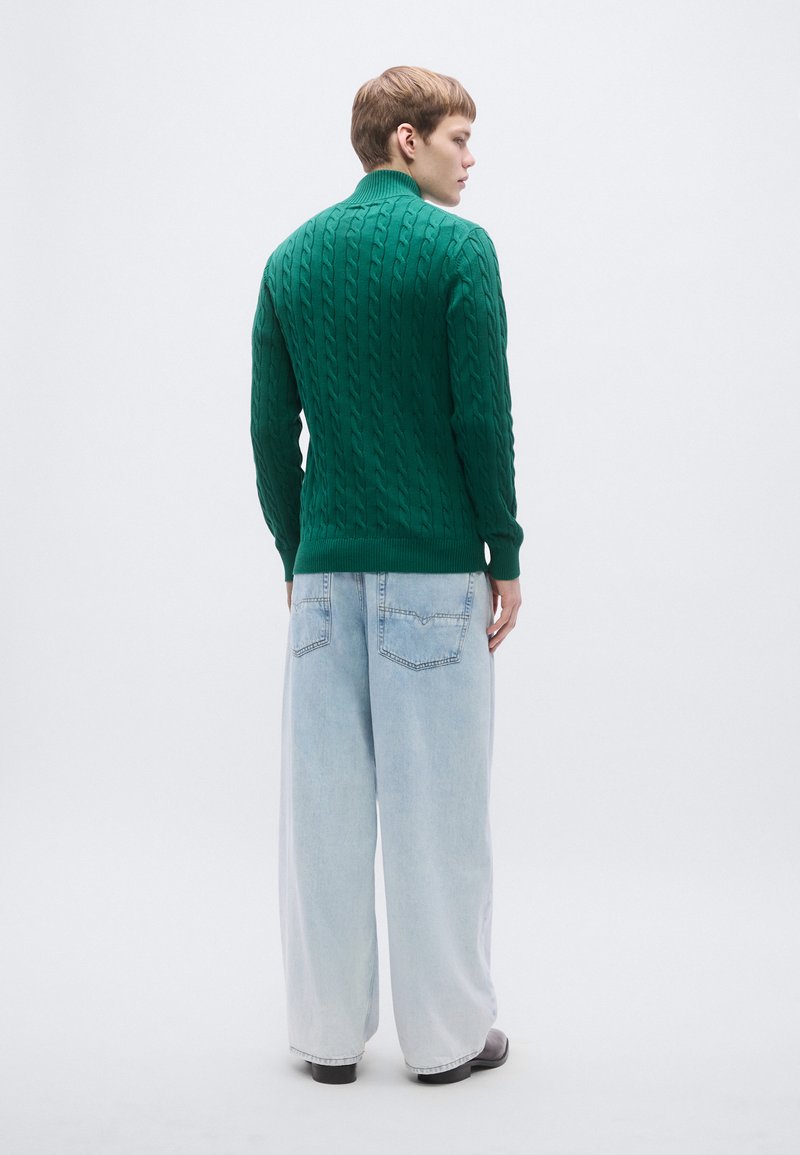 Green cable-knit sweater with a high collar, paired with light blue wide-leg jeans. The fabric has a textured pattern with ribbed cuffs.