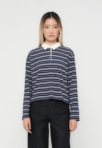 Marine stripete langermet poloskjorte med hvit krage og knappelukking med tre knapper, sammen med mørkejeans. Teksturen virker jevn.