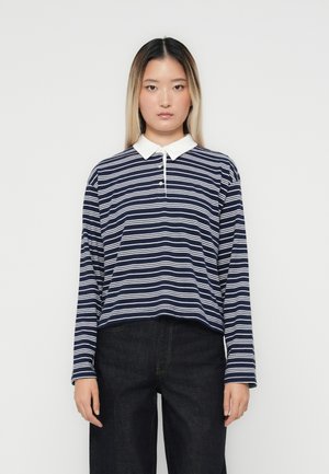 Polo a maniche lunghe a righe blu navy con colletto bianco e patta con tre bottoni, abbinato a jeans scuri. La texture appare liscia.