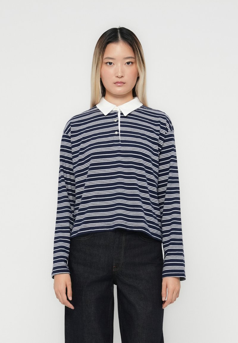 Marine stripete langermet poloskjorte med hvit krage og knappelukking med tre knapper, sammen med mørkejeans. Teksturen virker jevn.