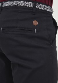 Black pants with a textured belt featuring a geometric pattern. Leather logo patch on the rear pocket, contrast stitching detail.