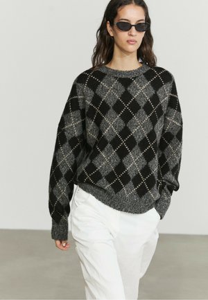 Frau trägt schwarzen und grauen Argyle-Pullover, weiße Hose und schwarze Sonnenbrille, geht mit einer Hand in der Tasche vor einfarbigem Hintergrund.