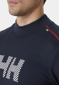 Navyblauwe trui met hoge hals, voorzien van een wit gestreept logo-ontwerp op de borst en rode accentstiksels op de schouders. Zachte stofstructuur.