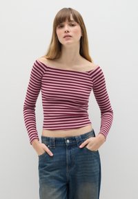 Gestreiftes Off-Shoulder-Top in Maroon und Pink mit horizontalen Streifen, kombiniert mit hochgeschnittenen blauen Jeans. Das Model posiert mit den Händen in den Taschen.
