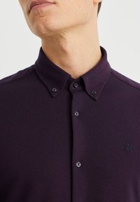 Tumma purppurainen kauluksellinen paita, jossa on napillinen etuosa, teksturoitu kangas ja pieni kirjailtu lintukuvio.