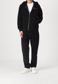 Svart zip-up hoodie med dragsko, framficka och logodetalj. Tillsammans med svarta löst sittande byxor och vita sneakers.