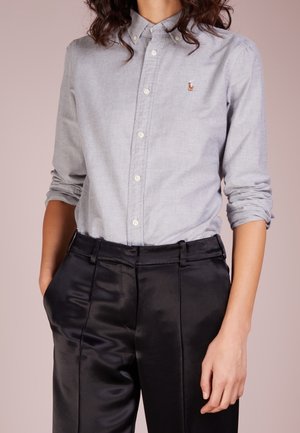 Kobieta ubrana w jasnoszarą koszulę na guziki z podwiniętymi rękawami i czarne satynowe spodnie, stojąca z ręką w kieszeni.