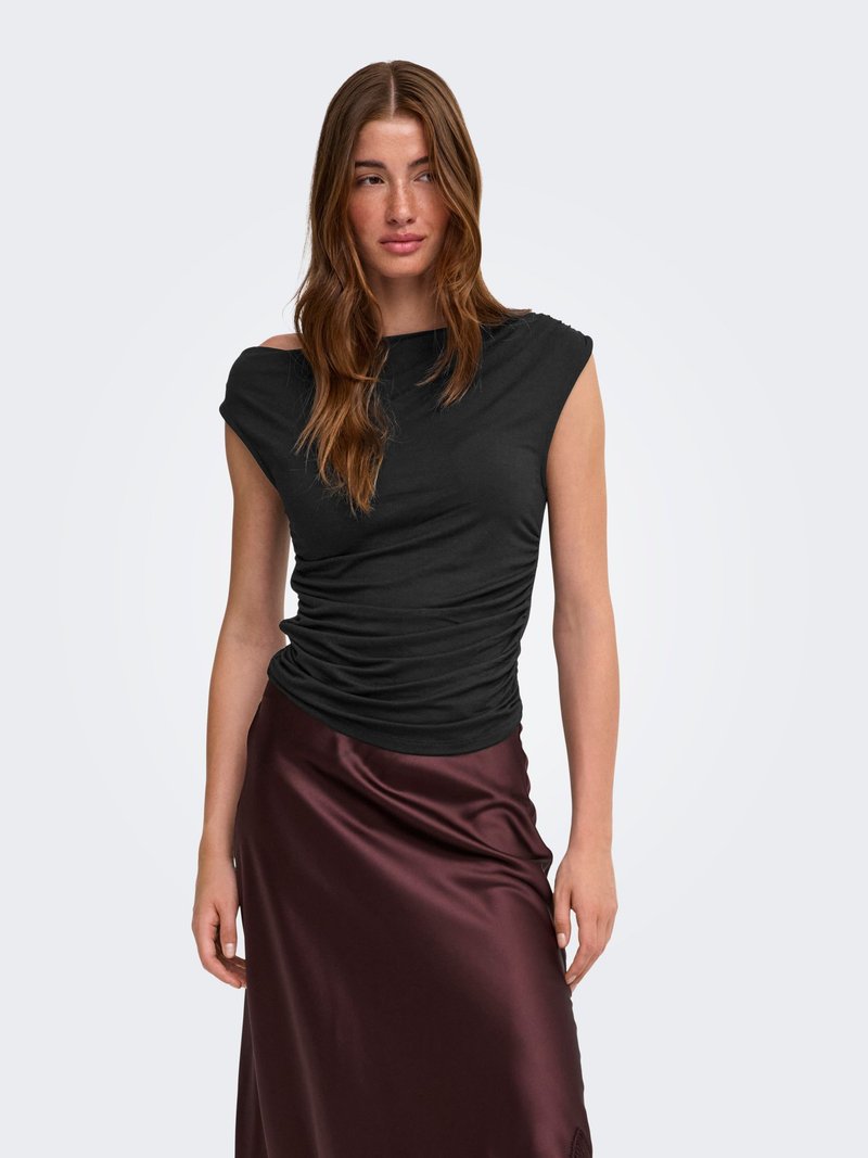 Femme aux longs cheveux bruns portant un haut noir sans manches froncé et une jupe en satin brun foncé brillante sur un fond clair uni.