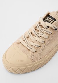 Beige canvas sneaker with rubber toe cap and textured sole, featuring cream laces and branding on the tongue and lace sides.