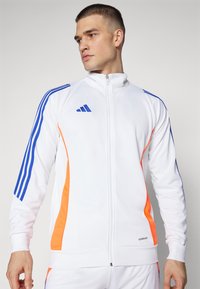 Hvid lynlåsjakke med blå og orange detaljer, der har tre blå stribede linjer på ærmerne og "AEROREADY" mærke ved sømmen.