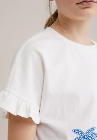 Camiseta de algodón blanca con mangas cortas onduladas y detalle de mariposa bordado azul en la parte delantera, con escote redondo.
