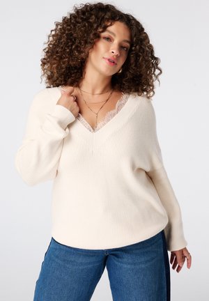 Vrouw met krullend haar die een crèmekleurige V-hals trui draagt over een kanten top en blauwe spijkerbroek, poseert met één hand bij haar kraag en de andere ontspannen.