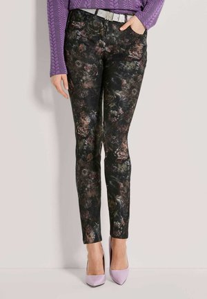 Frau trägt schwarze Hose mit Blumenmuster, lila Zopfpullover, weißen Gürtel und lavendelfarbene High Heels, steht mit einer Hand in der Tasche.