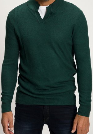 Homme portant un pull en maille à manches longues vert foncé avec un décolleté en V, par-dessus un t-shirt blanc et un jean foncé.