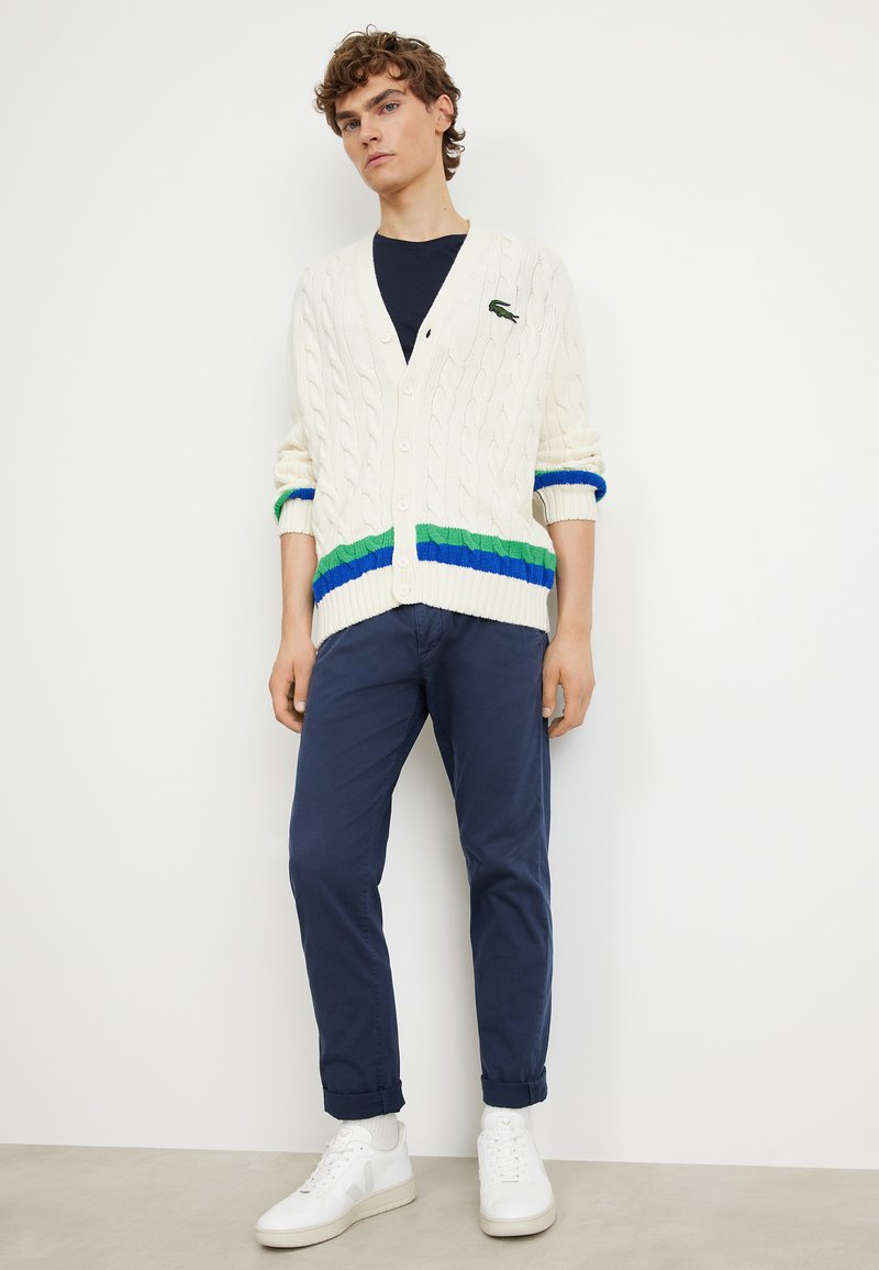 Gilet en maille torsadée crème avec des rayures bleues, vertes et navy aux poignets et à l'ourlet, associé à un chino navy et des baskets blanches.