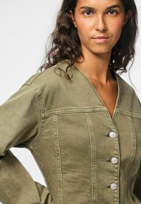 Donna con capelli ricci scuri che indossa una giacca verde oliva con bottoni, in piedi con una mano sulla vita, sfondo neutro.