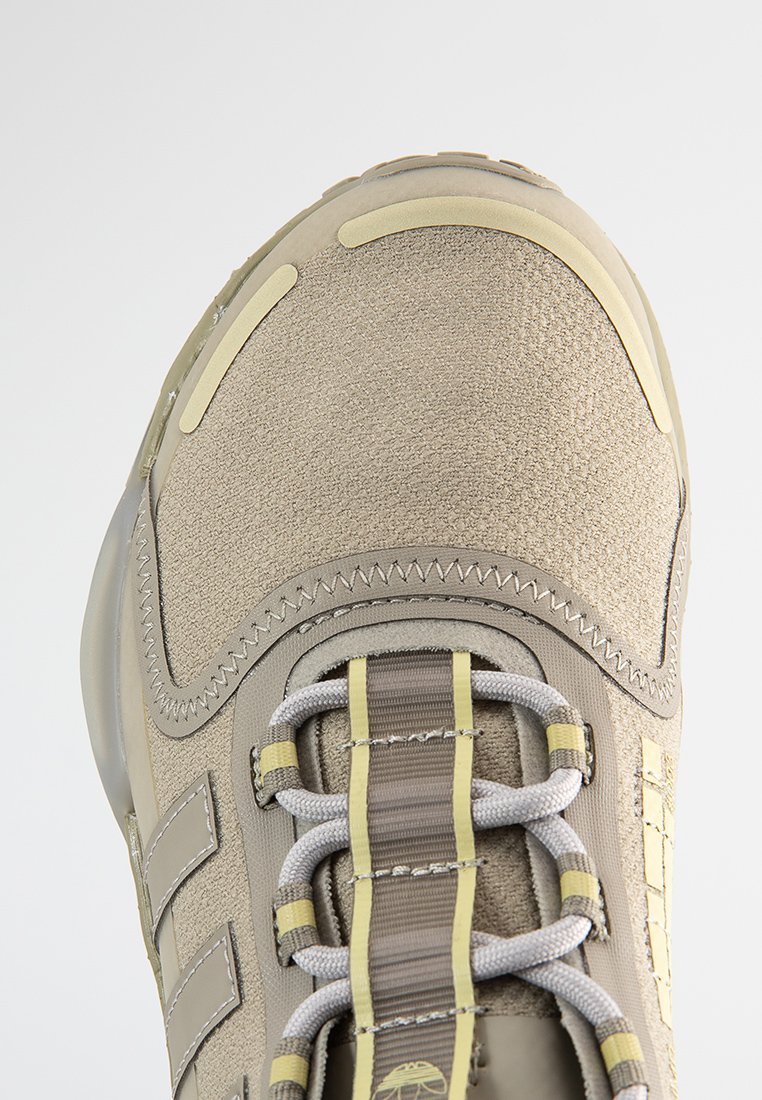 Zapatilla deportiva beige con una parte superior texturizada, suela de goma, puntera reforzada y cordones grises en contraste. Detalles amarillos a lo largo del costado.