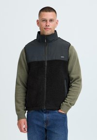 Gilet in pile nero con colletto alto, chiusura con zip e maniche lunghe verde oliva. Presenta una texture morbida e materiali a contrasto.