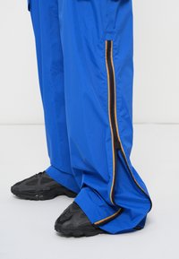 Calças leves azuis com detalhe de zíper lateral preto e amarelo, acompanhadas de ténis pretos texturizados. O material parece suave e durável.