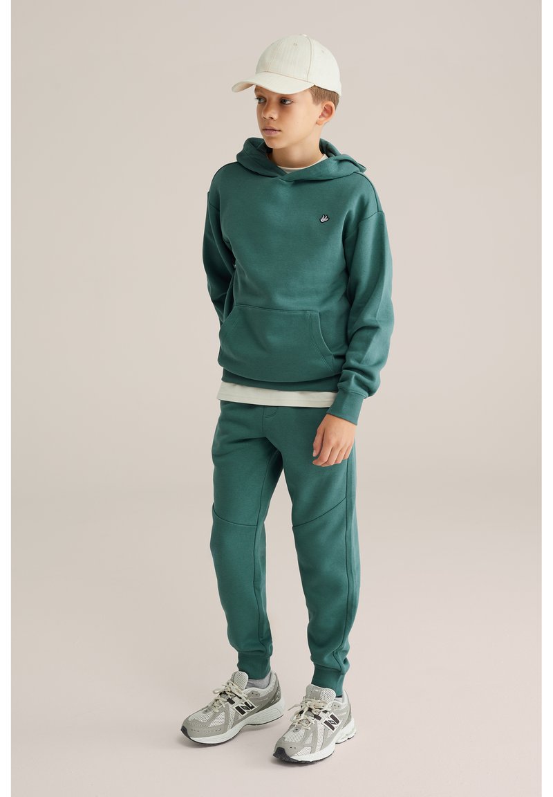 Junge trägt einen türkisfarbenen Hoodie und passende Jogginghosen, weiße Mütze und graue New Balance Sneaker, steht vor einem einfarbigen hellen Hintergrund.