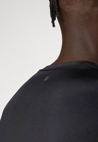 Fekete sportos póló sima textúrával és testhezálló szabással. Kerek nyakú, és finom márkajelzés található a hátsó vállrészen.