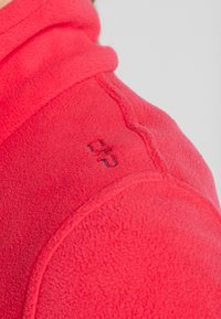 Gros plan d'une veste polaire rouge montrant une couture d'épaule et un petit logo en relief sur un tissu doux et texturé.