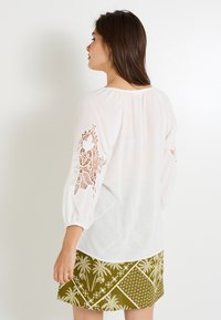 Blusa blanca de manga larga abullonada con intrincados patrones de encaje troquelado. Combinada con una falda verde adornada con motivos de palmeras.