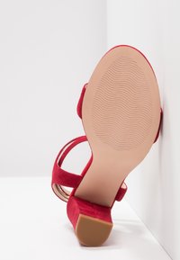 Sandale à talons hauts en daim rouge avec brides autour de la cheville, dotée d'une semelle extérieure en caoutchouc beige texturée et d'un talon carré.