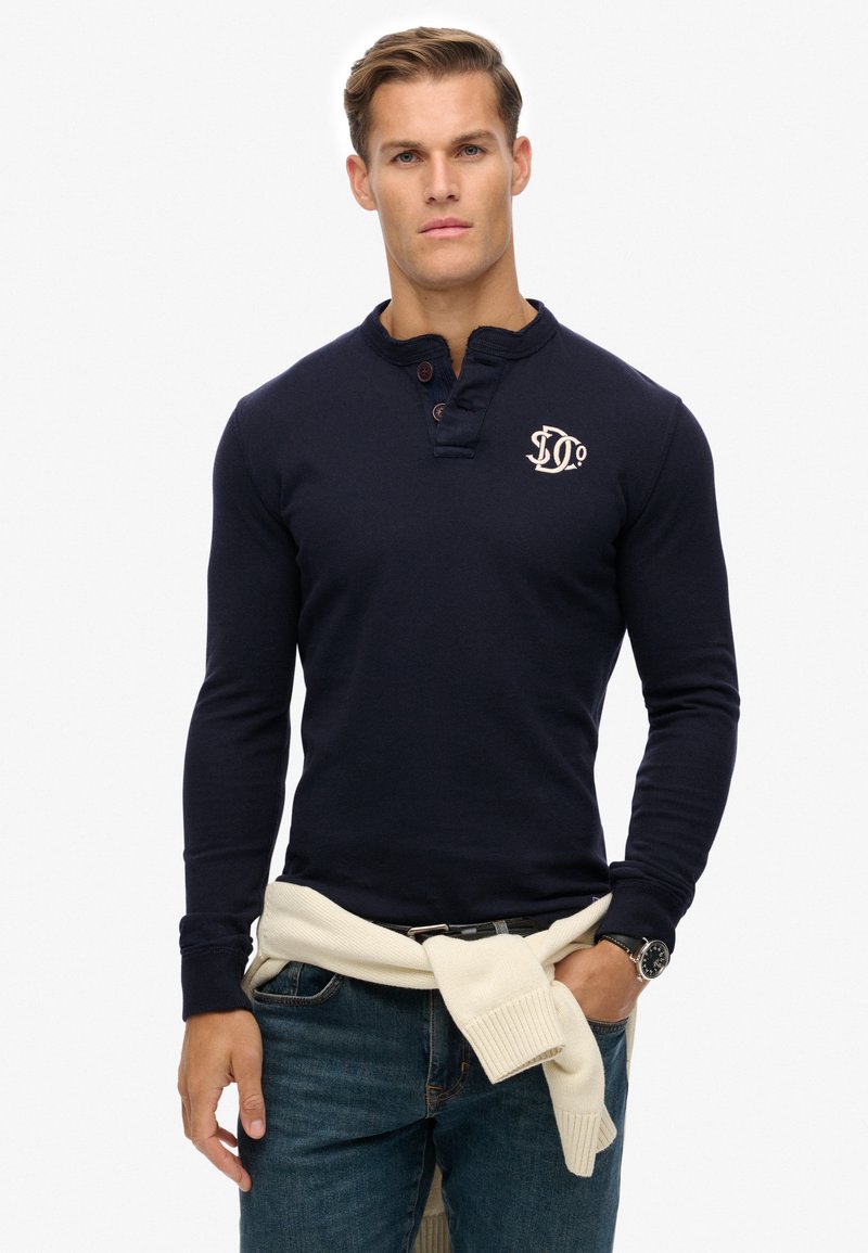Marineblaues Langarm-Henley-Shirt mit geknöpfter Blende, gerippten Bündchen und einem gestickten Logo. Getragen mit einem hellen Pullover, der an der Taille gebunden ist.